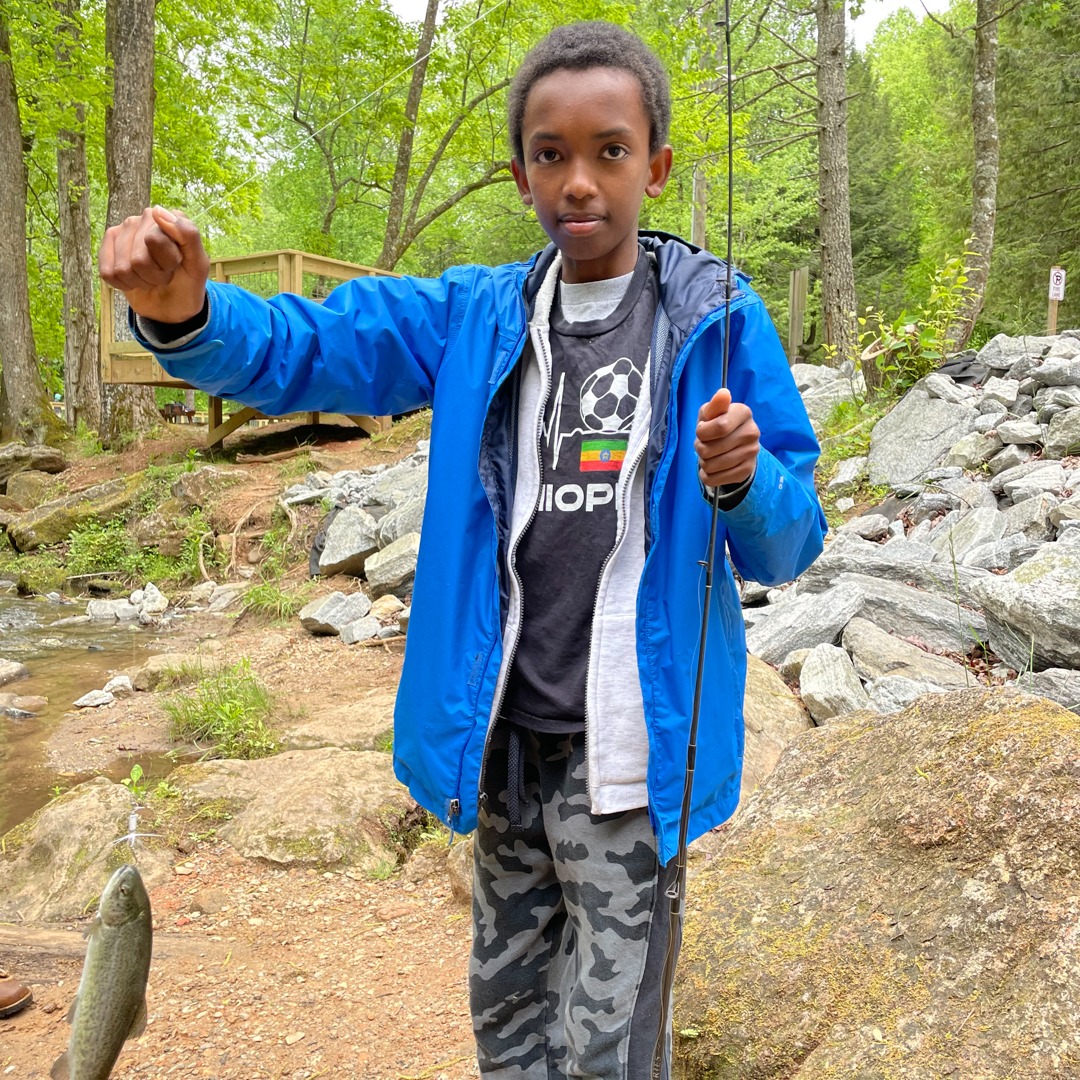 A young boy in a bright blue jacket holds a recently caught fish
