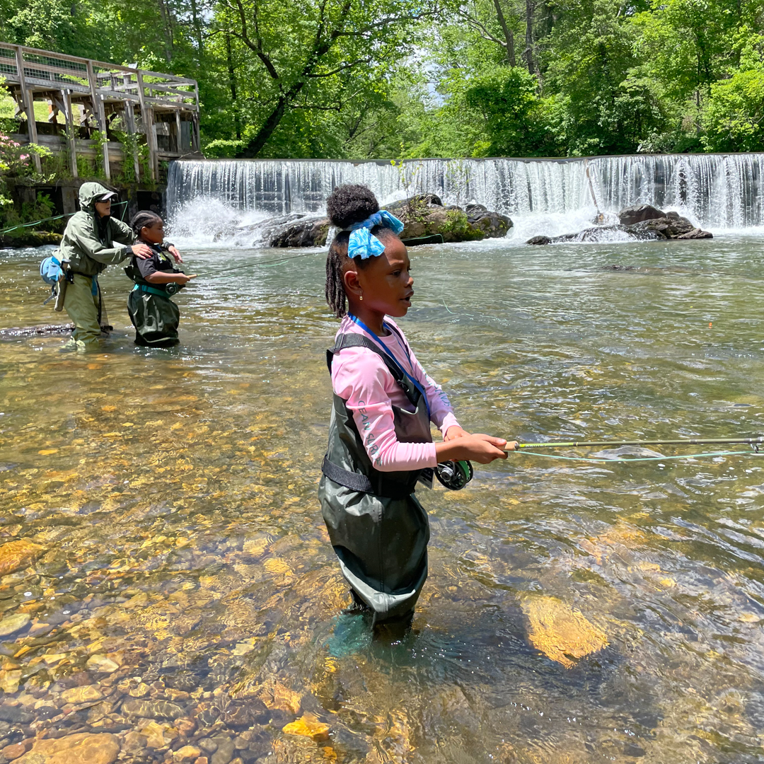 A little girl in a pink shirt and waders stands in a stream reeling a fishing pole with an instructor helping another child fish in the background