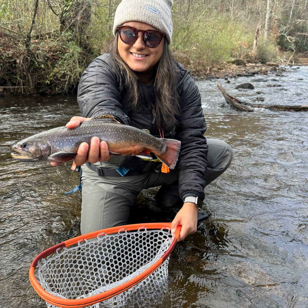 Woman wearing a gray beanie and suglasses kneels in a stream holding a brook trout she caught with the net in her other hand