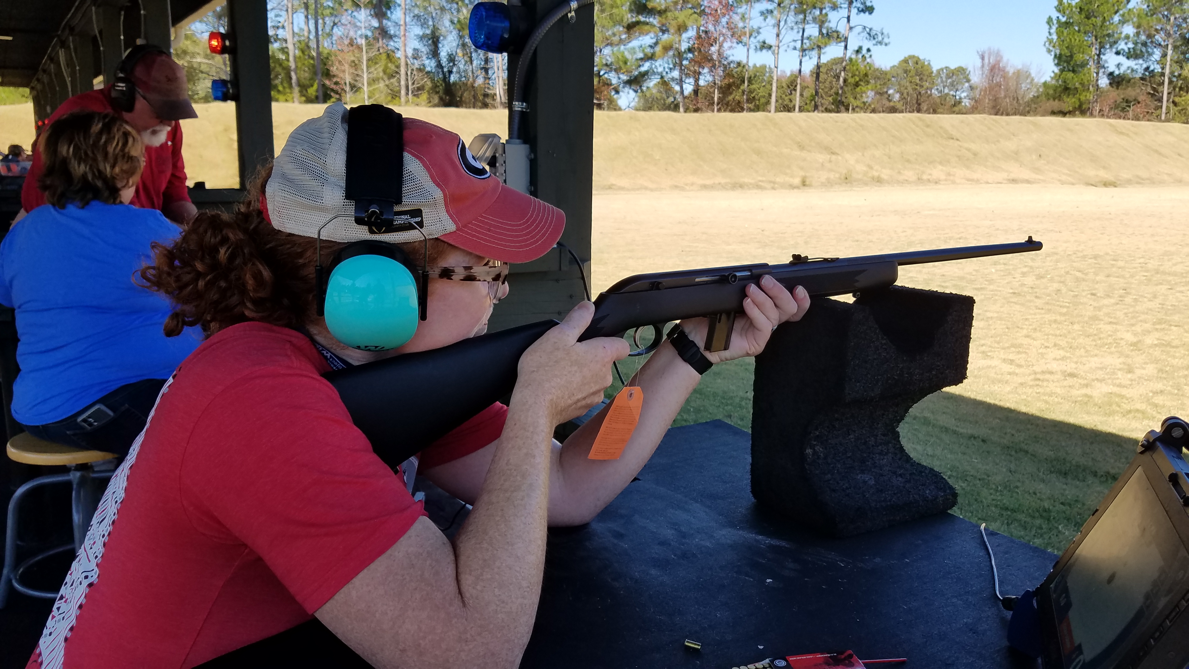 Woman swith red hair and a red hat and t-shirt sits mounted at a shooting stand, aiming a rfile.