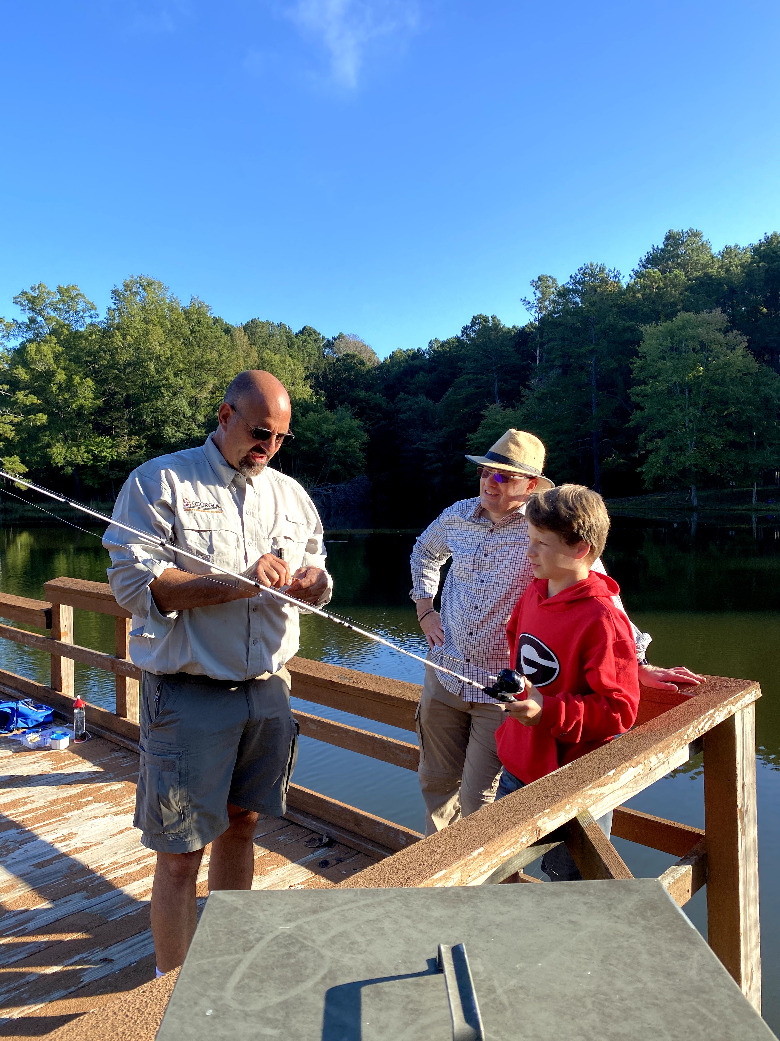 A boy and two adult men stand on a dock learning how to attach a fishing hook onto a pole.