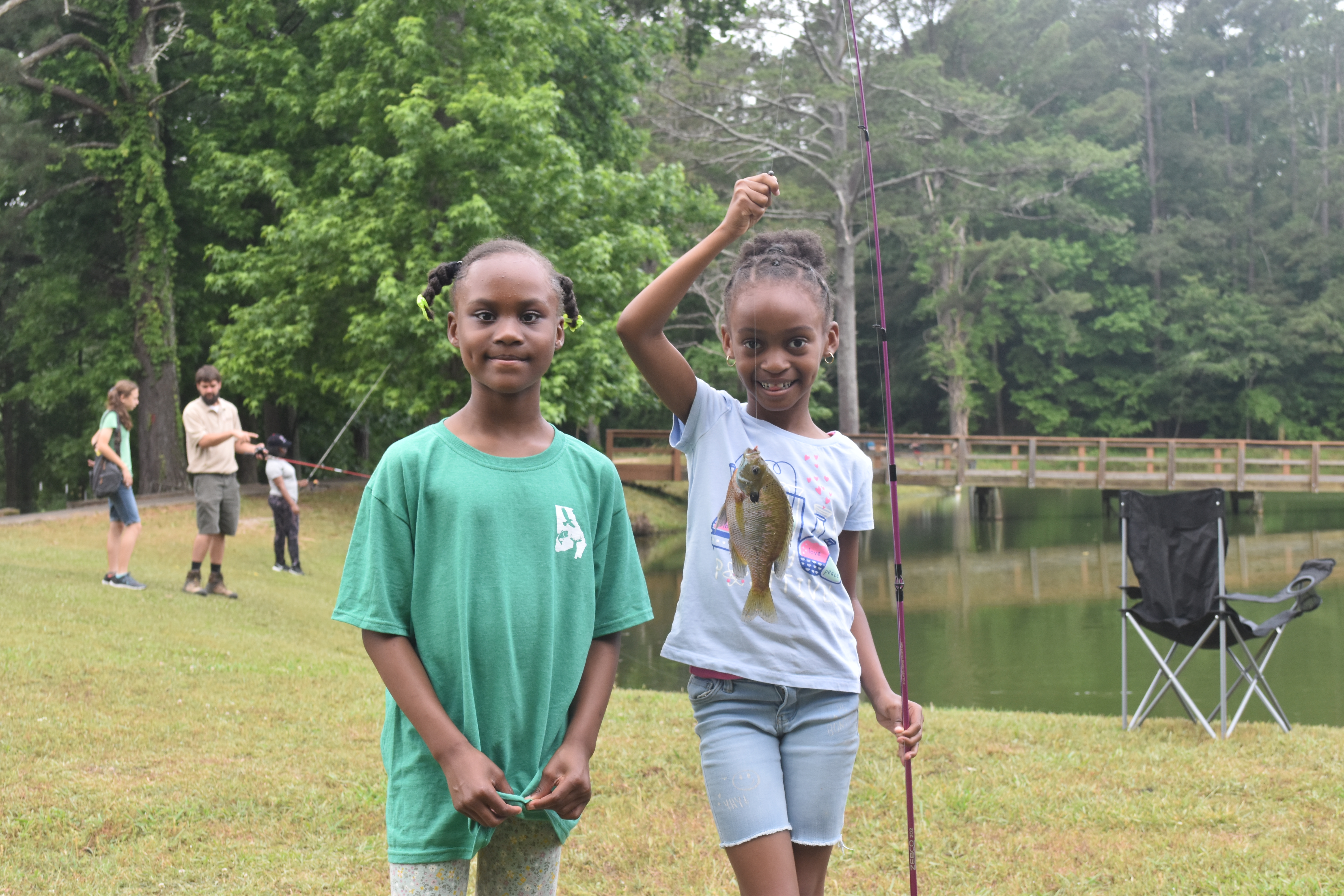 Two young girls pose in front of a lake showing off a fish they caught.