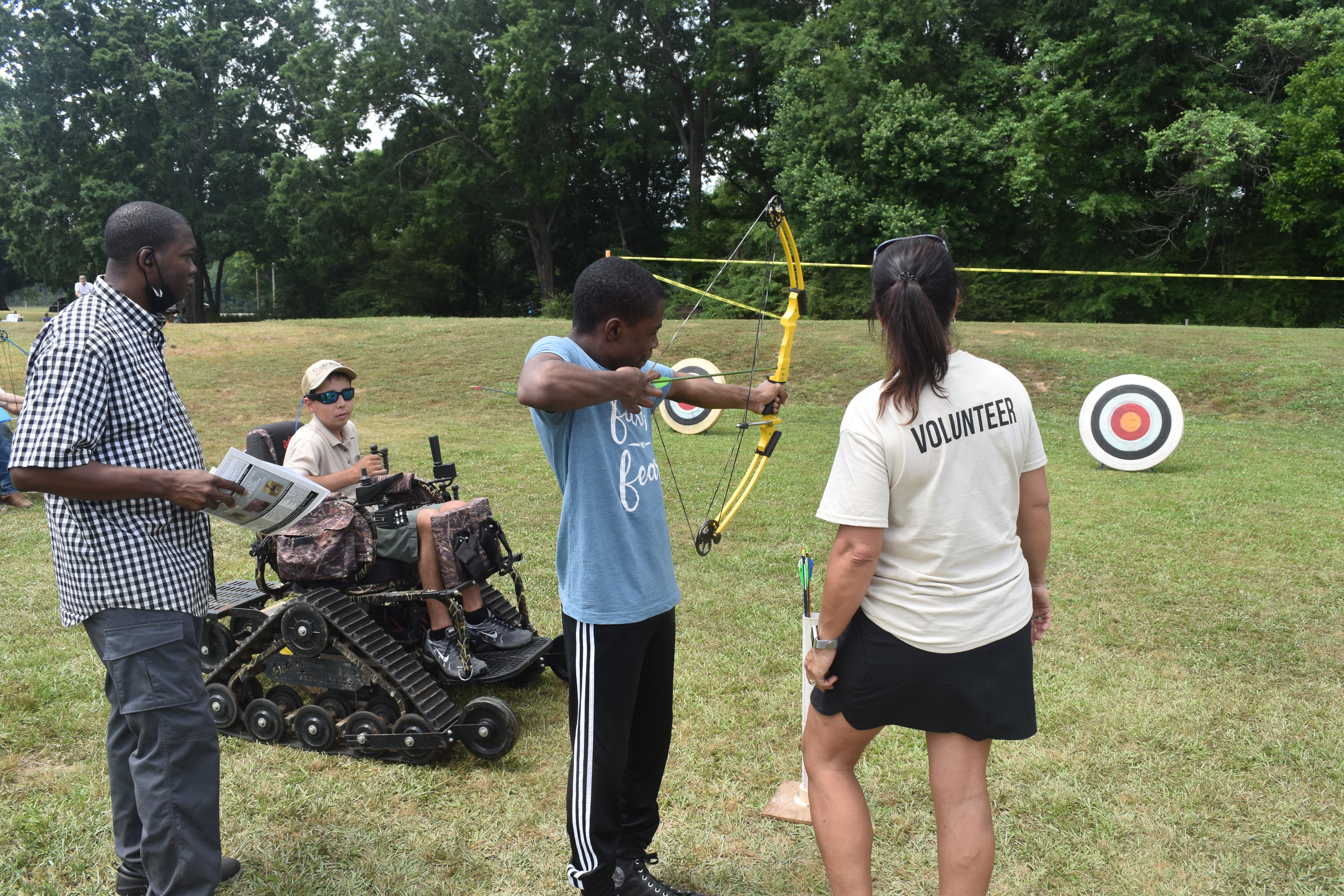 A child is being instructed on how to shoot a bow and arrow, a boy in an off-terrain mobility scooter rides next to him.