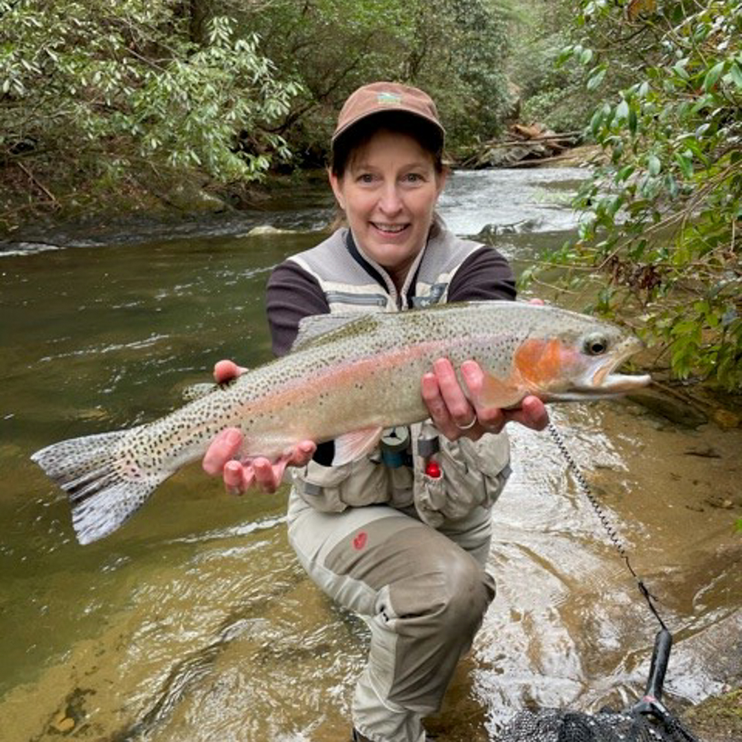 A woman in a red hat kneels on one knee in a stream presenting a large rainbow trout.
