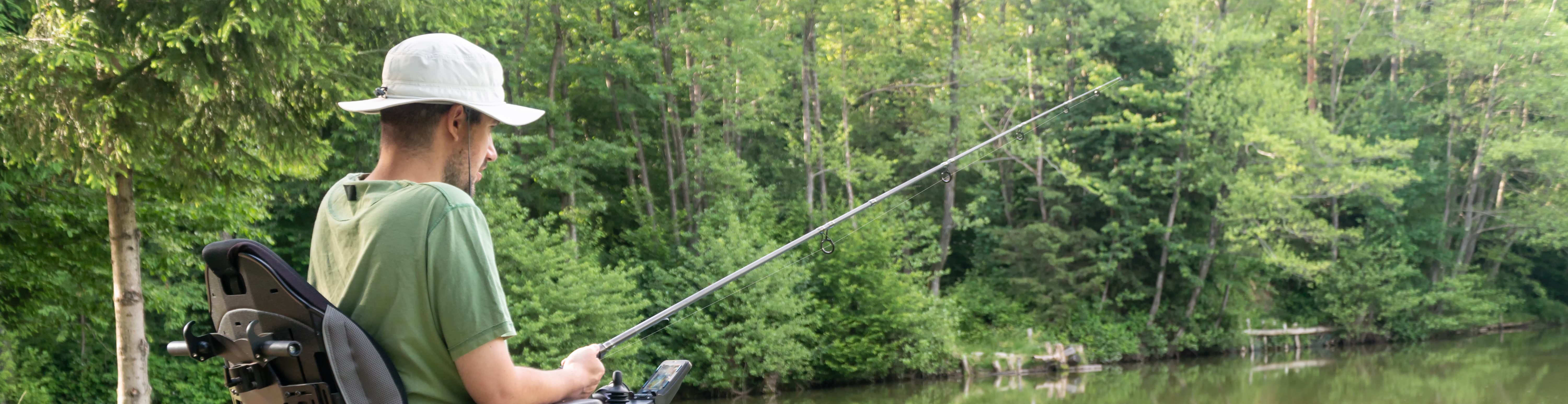 A man in a wheelchair casts a fishing pole on the bank of a lake.