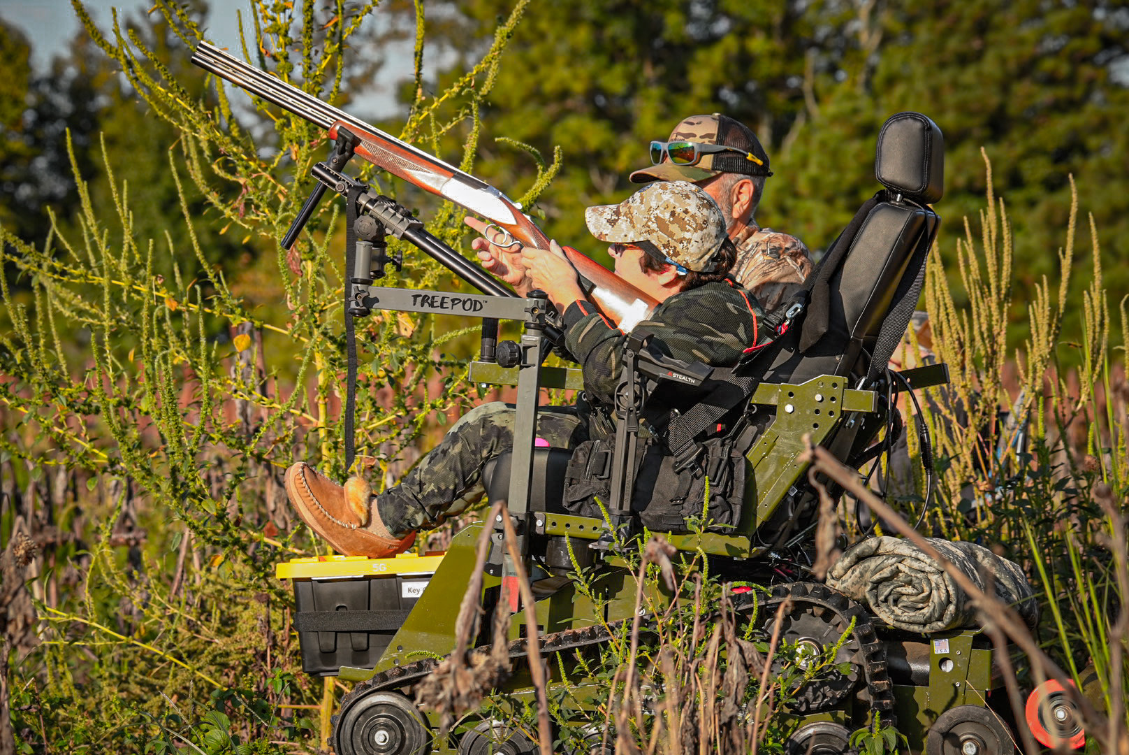 A female hunter dressed in camo sits in an all-terrain track chair with her feet propped up on a black box. She has a shotgun in a supportive rest while scanning the sky for doves out in a field. Beside her sits a male mentor also in camo assisting her with steadying the potential shot.