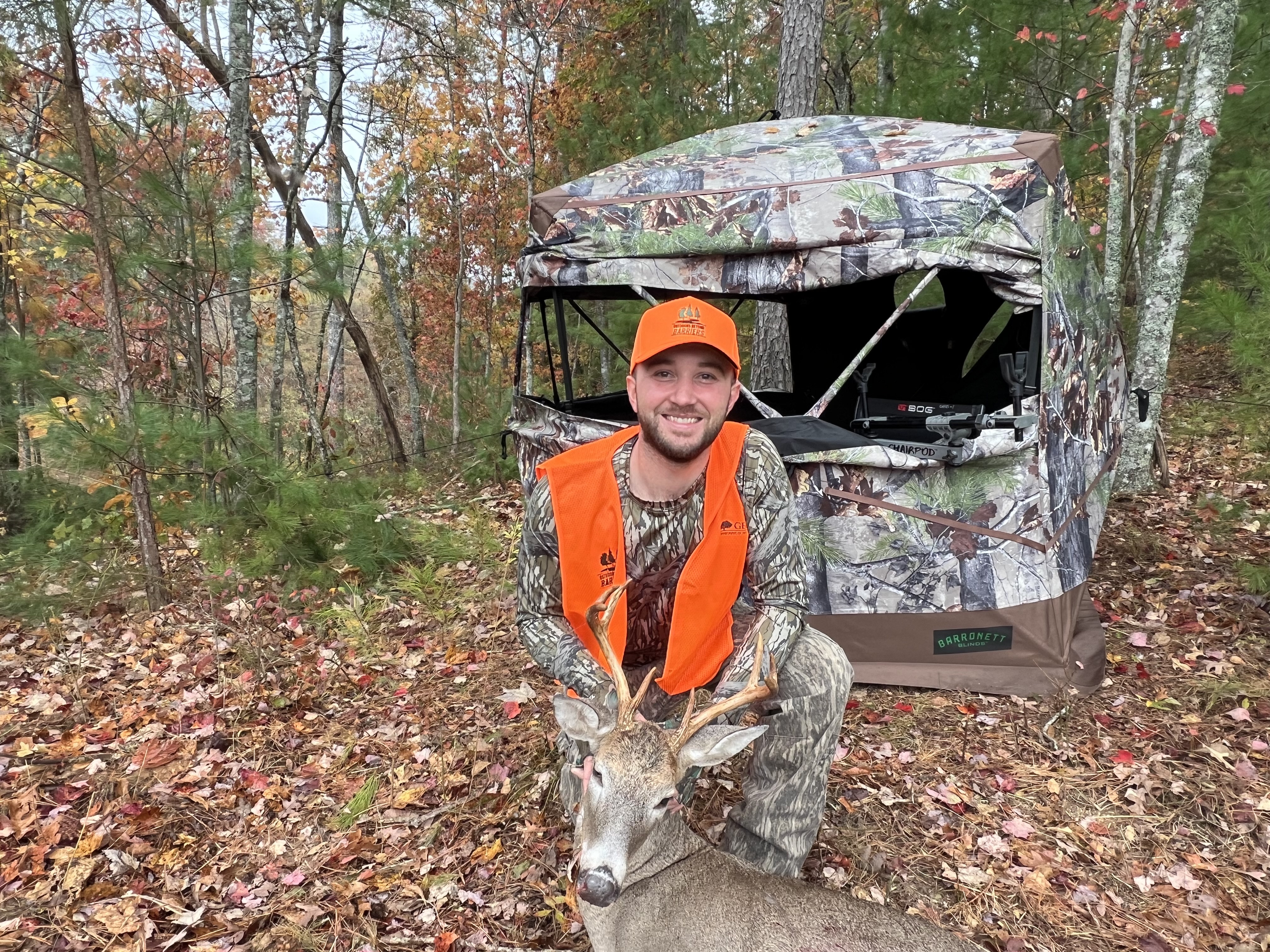 A male hunter in camo and hunter orange kneels on the ground smiling with his 8-point buck he harvested in front of the blind he had been sitting in; adaptive hunting equipment can be seen coming out of the blind’s window.