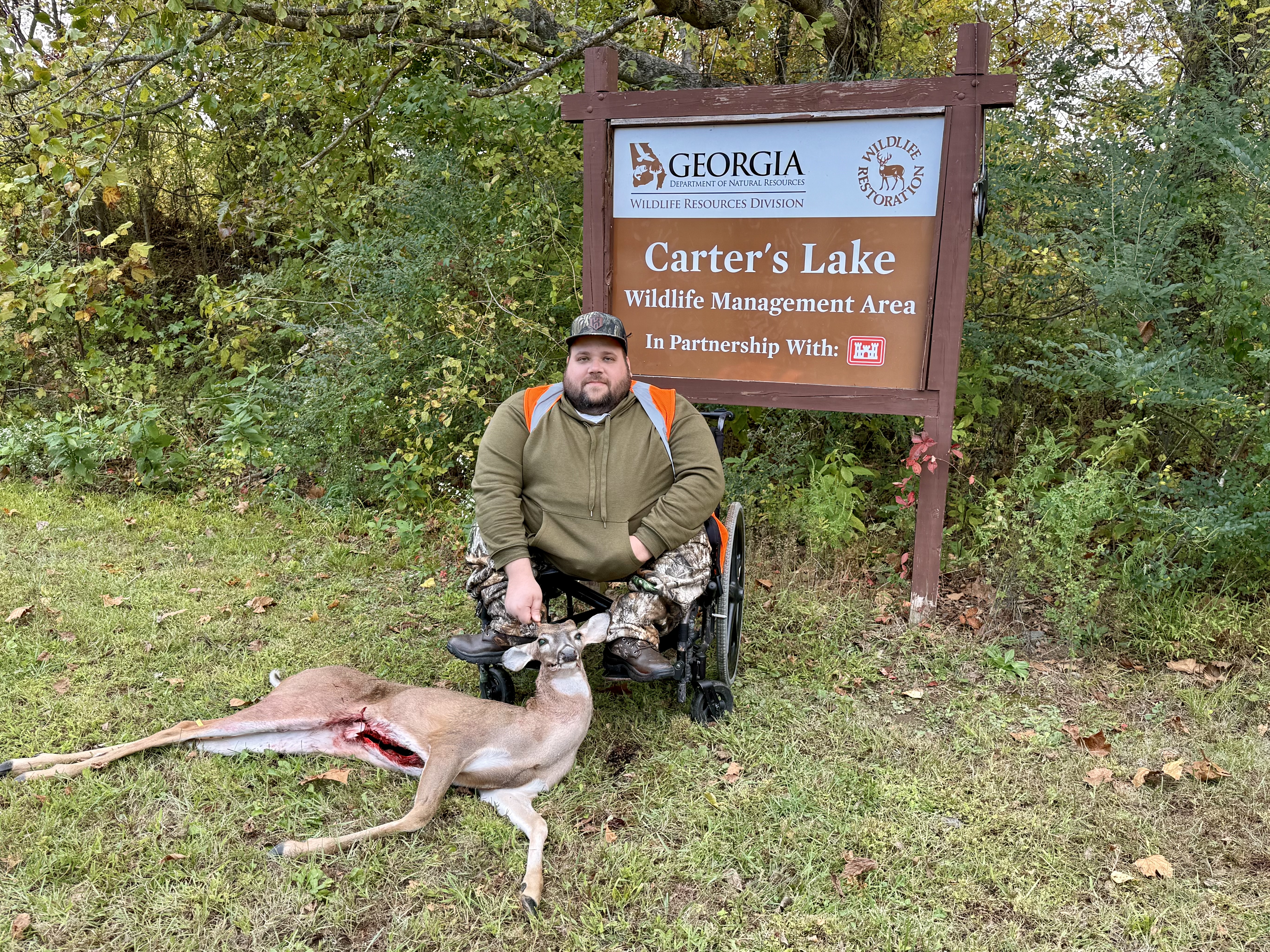 A male hunter sits in his wheelchair smiling in camo along with his harvested spike buck in front of the Carter’s Lake Wildlife Management Area’s sign.