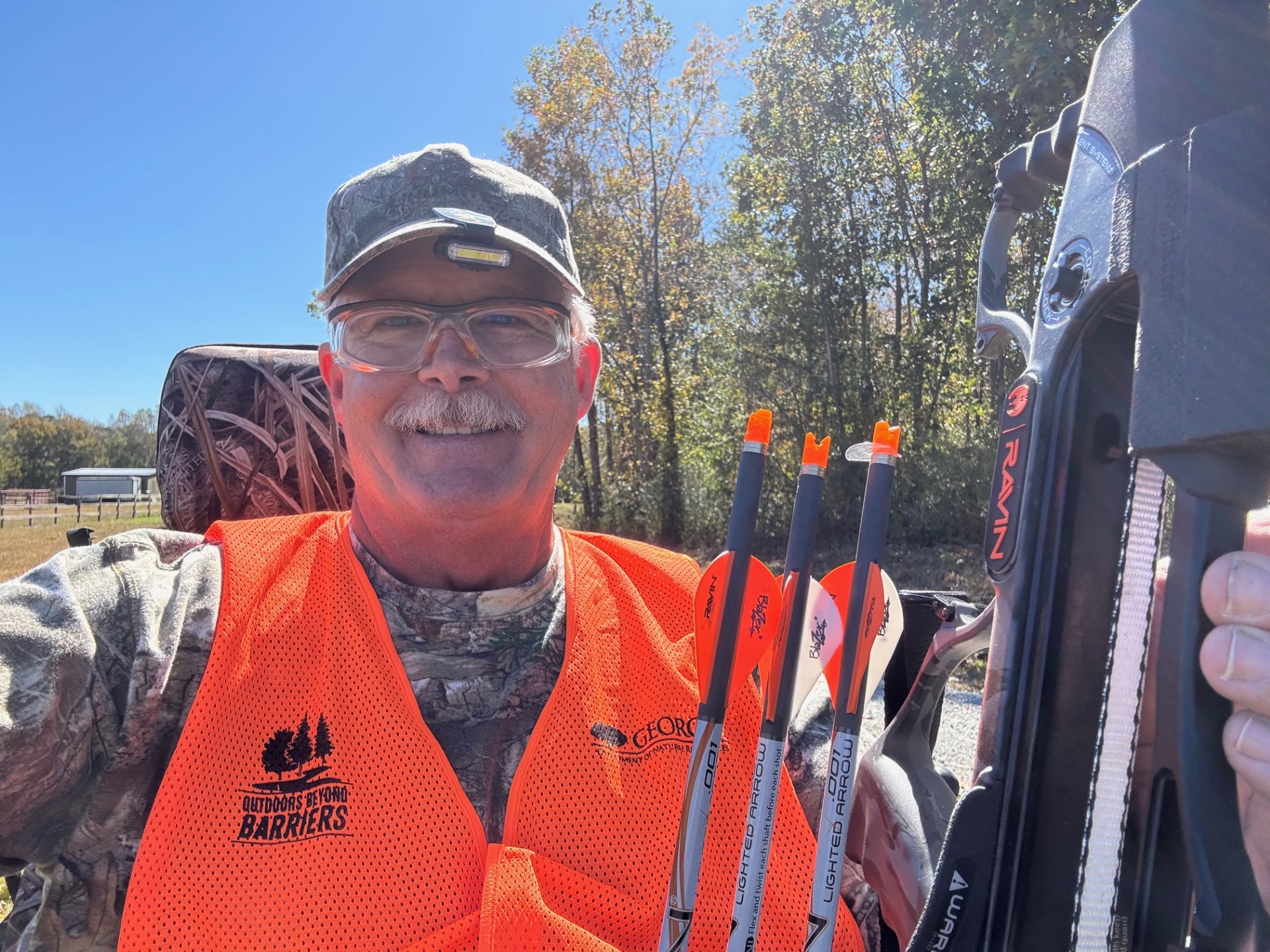 A smiling man with a mustache in an Outdoors Beyond Barriers hunter orange vest, camo and safety glasses holds his crossbow while sitting in his all-terrain chair in an outdoor setting.